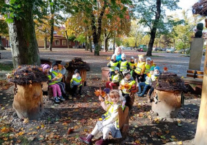 Grupa dzieci siedzi na ławeczkach i pieńkach drewnianych obok pomnika T. Kościuszki.